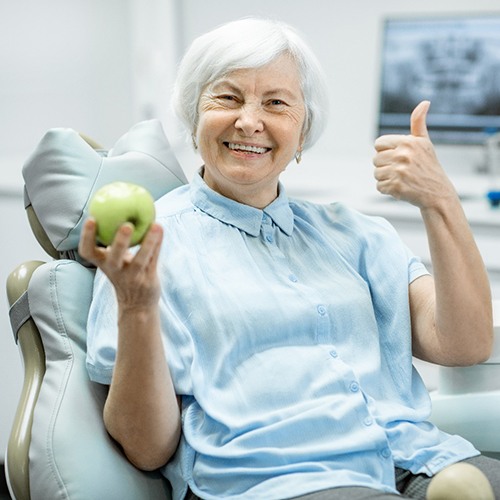 elderly woman giving a thumbs-up and holding an apple in the dental chair 
